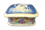 LIMOGES -  Caixa confeccionada em porcelana francesa sec. XX, esmaltada e policromada com decoração floral ao redor, e com a tampa com cena romântica de casal nas nuvens sob as estrelas, em relevo; Medindo 5 x 10 cm
