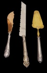 Três peças de servir, incluindo faca e espátulas, com cabos confeccionados em prata ricamente ornamentada, e partes funcionais em metal (com acabamento prateado e dourado). Medidas: 29,8 cm, 37 cm e 26,5 cm.