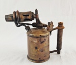 Antigo maçarico a querosene em metal, marca Primus. Corpo cilíndrico com reservatório inferior, empunhadura lateral com mola e conjunto de queima com bico alongado. Peça com pátina natural do tempo. Medida: 23 cm de altura.
