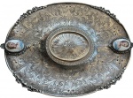 PRATA. Elegante e imponente bandeja / centro de mesa em prata 833 contrastada, de provável manufatura portuguesa, datada do início do século XX, apresentando exuberante trabalho de cinzelagem em alto-relevo com rica decoração em arabescos vegetalistas, sobre fundo pontilhado, parte central é marcado por medalhão circular em relevo radial e aplicações decorativas e dois delicados medalhões ovais em porcelana pintada à mão, representando bustos femininos de inspiração clássica, alças laterais trabalhadas. Peça contrastada PRATA 833, típica da prataria portuguesa do período, tendo sido comercializada por tradicional casa do ramo, atribuída à Casa Castro, importante revendedora de peças finas no Brasil no século XX. Medidas: 32 x 23 cm. Peso: 601 g