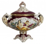 ITÁLIA - Imponente centro de mesa em porcelana italiana de Nápoles, ricamente decorada em relevo, com cenas clássicas policromadas, figuras aplicadas nas alças e abundante douração. Tampa original, base pedestal e esmaltação em tom vinho, típica da produção napolitana do século XIX. Peça ornamental de forte impacto decorativo e excelente presença. Alças em folhagens com figuras de querubins. Registro na base. Mede 40 x 35 cm.