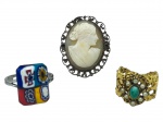 BIJUTERIAS, lote com três (3) peças, sendo: A) anel em metal prateado, com aplicação quadrangular em vidro de Murano, aro ajustável, sinais de uso, B) 1 broche/pingente em metal dourado, com camafeu oval representando busto feminino, emoldurado por trabalho vazado, medindo 3x2,5cm e C) anel em metal dourado, com pedra verde central e pequenas contas peroladas, aro ajustável, peças com sinais de uso, vendidas no estado da foto.