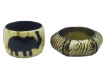 BIJUTERIAS, dois (2) braceletes, sendo: A) 1 confeccionado em resina imitando marfim, decorado por figuras de elefantes na cor preta, apresenta bicado na borda, medindo 7cm e B) 1  com estrutura em metal dourado, incrustado por placas em resina com pintura tigrada, medindo 9,5x6,5cm, sinais de uso.