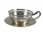 METAL ALPACA , Uma (1) xícara com pires, em metal espessurado à prata, ricamente trabalhada, apresentando bordo vazado em desenho geométrico, frisos em relevo, bojo com decoração cinzelada e alça em elegante curvatura. Medida : 4 x 12 x 9 cm , falta o recipiente interno