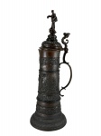 ESTANHO, um (1) grande tankard confeccionado em estanho com patina na tonalidade bronze, ricamente cinzelada por elementos vegetalistas e alça curva encimada por figura angelical e pega da tampa representando figura masculina, medindo 48cm de altura, apresenta leve mossa na borda.