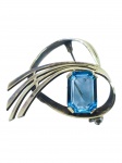 BIJUTERIA FINA, um (1) broche, confeccionado em metal dourado, cinzelado por elementos vazados e estilizados, com incrustação de pedra na tonalidade azul, possivelmente água marinha, medindo 4,5x4cm.