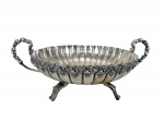 METAL PRATEADO, uma (1) fruteira/centro de mesa confeccionado em metal espessurado a prata, corpo em gomos e borda cinzelada em cachos de uva e folhas de parreira, apoiada sobre 3 pés recortados e alças laterias vazadas, apresentam falhas na prateação, medindo 17x40x30cm.