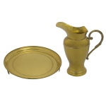 Conjunto de jarra e salva em metal dourado liso, ornado por frisos. Bojo de formato balaústre com torsade à meia altura, encimado por largo bico revirado e ladeado por alça lavrada em S. Base circular em degraus salientes. Art Prata. 26 x 23 cm. Salva com borda alta, realçada por filetes em debrum, centralizando fundo liso decorado por faixa geométrica, apoiado em pés retos.3 x 28 cm.