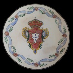 Prato em faiança portuguesa, ornamentado em policromia com as armas do Império português encimadas por coroa. Borda realçada por guirlanda com fitas e florões. Marca do verso não registrada. 3 x 39 cm.