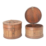 Par de caixas circulares com tampas reclináveis, construídas em madeira clara e lisa. Apresentam laterais trançadas em junco. Tampas no estilo, com cintas transversais e alças superiores. 20 x 24 cm.