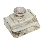 Tinteiro alemão em cristal transparente, estilo Art Déco, de formato quadrangular. Frente rebaixada e laterais com porta canetas escavadas. Apresenta ao centro recipiente cilíndrico com tampa em prata, ornada por filete e ramos floridos. Contraste da prata alemã de teor 800 mls, usado a partir de 1888 e marca do prateiro. 6 x 9 x 7 cm.