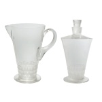 LALIQUE FRANCE  Conjunto em cristal transparente, constando de uma jarra cônica, lapidada e ornada na parte inferior por 3 frisos com motivos triangulares, e encimada por gargalo e tampa no estilo. Base circular lisa. Acompanha jarra moldada e ornamentada no mesmo modelo, ladeado par bico curto e alça em asa. Marcas gravadas da Cristallerie. 21 x 20 cm (jarra) e 24 x 12 cm (garrafa).