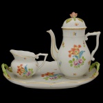 HEREND  Elegante conjunto em porcelana húngara, ornamentado em policromia floral e filetes a ouro. Constando de bule balaústre para café, leiteira bojuda e bandeja oval recortada. Apresentam bordas onduladas, bicos recurvos, alças em asa e tampa semiesférica encimada por rosa. Pegas da bandeja em meia argolas torcidas. Marcas da manufatura sob as bases.  33 x 26 cm (bandeja). 12 x 18 cm (bule) e 9 x 13 cm (açucareiro).