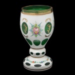 Copo em cristal da Boêmia de cor verde esmeralda, realçado por adornos a ouro, revestido por overlay leitoso, lapidado com janelas ovaladas. Apresenta decoração com buquês policromados. Bojo de formato cônico com borda lisa, apoiado em haste anelada sobre base semiesférica decorada por guirlanda florida.16 x 9 cm.