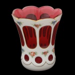 Copo em cristal da Boêmia de formato cônico levemente abaulado, de cor rubi, realçado por filetes a ouro. Apresenta revestimento com overlay leitoso, lapidado com janelas ovaladas e realçado por buquês policromados. Bojo de formato cônico com borda lisa, apoiado em haste anelada sobre base semiesférica decorada por guirlanda florida. Base arredondada. 11 x 10 cm.