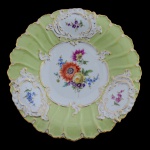 MEISSEN  Belíssimo prato em porcelana alemã, policromada e dourada, com borda ondulada. Apresenta margem moldada com folhas alinhadas em leve relevo, intercaladas por reservas ovaladas com ramos floridos e volutas. Ao centro, composição floral emoldurada por volutas. Marca da manufatura sob a base. 30 cm.