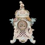 Alemanha. Séc. XIX - Delicado relógio de mesa em porcelana alemã policromada e dourada, moldada em alto relevo. Caixa retangular montada com treliças a frente, ladeada por putti musicistas, flores e folhagens. Centro com mostrador circular e assinatura do relojoeiro Louis Martin (1823-1894). Verso liso decorado por arranjos florais. Tampa abobadada, acompanhando o estilo com vazados e florões, encimada por anjinho. Base apoiada em volutas laterais, centralizando reserva com pássaros. Marca da manufatura sob a base. 50 x 34 x 18 cm.