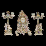 Alemanha. Séc. XIX - Exuberante Garniture de Cheminée, em porcelana alemã policromada e dourada. Apresenta relógio de mesa, de formato alongado, profusamente ornamentado em alto relevo com treliças, folhas, flores e Putti distribuídos sobre a frente do relógio. Ao centro, mostrador circular de fundo branco com a marca do relojoeiro. Verso liso decorado por arranjos florais. Base apoiada em volutas assimétricas, centralizando estilizações onduladas. Acompanha par de candelabros para 2 velas, construídos no mesmo estilo, com colunas retas, encimadas por braços recurvos e ponteiras floridas. Bases apoiadas em pares de anjos sobre arabescos laterais. Marca da manufatura sob a base. 45 x 30 cm (relógio). 46 x 18 cm (candelabros). Acompanha chave e pêndulo original.