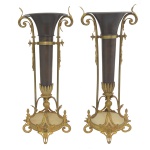 Original par de floreiras em metal preto com montagens em bronze polido. Bojos cilíndricos e alongados com bordas reviradas em cornetas, montadas em suportes com hastes laterais retas ornadas por anelados a meia altura e encimadas por volutas. Bases com pequenos plateaux circulares, bordadas por ricos frisos realçados por ponteiras e apoiados em arabescos. 53 x 18 cm.