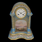 Relógio de mesa em porcelana policromada e dourada, de formato retangular. Apresenta frontão arqueado realçado por friso floral, ladeado por colunas em torsade. Centro com reserva ornada por paisagem lacustre encimada por mostrador circular em esmalte branco. Verso liso, decorado por composição com flores do campo. Base em degraus, com frente recortada e ornamentada por faixa com medalhões floridos. 35 x 27 x 17 cm.