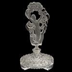 Belo perfumeiro em cristal transparente. Bojo cilíndrico e achatado, profusamente lapidado e ornado por facetas em xadrez, apoiado em pés recortados no estilo. Exuberante tampa acetinada e vazada, apresentando dançarina com véu realçado por flores e estilizações. 20 x 11 cm.