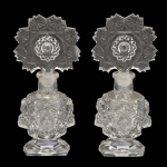 Par de luxuosos perfumeiros em cristal transparente. Bojos cilíndricos, ornamentados por máscaras e adornos em alto relevo. Bases chanfradas e esparramadas lisas. Tampas arqueadas, bordadas por estrelas centralizando medalhões com motivos raiados. 17 x 8 cm.