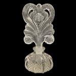 Original perfumeiro em cristal transparente. Bojo no formato de bolota dividida em gomos salientes, ornados por adornos em bico de jaca. Pequeno gargalo reto, encimado por exuberante tampa moldada e lapidada com volutas laterais sustentando, de cada lado, folhagens superpostas encimadas por grande coração vazado. 15 x 7 cm.