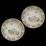 Par de pratos em porcelana, possivelmente alemã, com finas bordas onduladas. Margens vazadas por adornos florais repetidos e realçados a ouro. Apresentam ao centro composições policromadas com flores do campo. Sob as bases, marcas de manufatura não registrada.