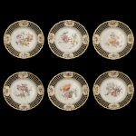 KPM  Conjunto de 6 pratos em porcelana alemã policromada e dourada. Apresentam finas bordas onduladas e margens vazadas em treliças realçadas por medalhões florais. Centros ornamentados por diferentes arranjos com flores do campo. Marcas da manufatura sob as bases.