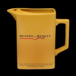 BENSON & HEDGENS - H C W Prompots  Made in Britain  Jarra para água  (água jug) em cerâmica de cor amarela. Produzida pelo Wade England para a marca de cigarros Benson & Hedgens. Na base a informação: TOBACCO SERIOUSLY DAMAGES HEALTH (O TABACO PREJUDICA SERIAMENTE A SAÚDE). No fundo marca da manufatura e as inscrições: H C W Prompots. 17 x 19 x 8 cm.