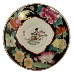 Prato em Porcelana Chinesa. Figura Feminina. C. 19501970 - Prato decorativo em porcelana chinesa, ricamente ornamentado no estilo Famille Rose. Apresenta figura feminina central em reserva, circundada por composição floral com peônias e flores estilizadas, executadas em policromia vibrante. Destaca-se pelo elegante fundo preto, enriquecido por delicada aplicação de douração, conferindo forte impacto visual à peça. 15 cm.