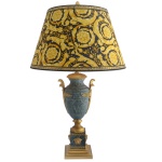 PAUL WUNDERLICH (German 1927-2010) For VERSACE. Modelo Barroco. Manufatura by Rosenthal - Luminária de mesa para 2 luzes, montada com urna de formato balaústre esculpida em mármore cinza rajado. Montagem ornamental em bronze dourado, com alças laterais reviradas e acopladas a folhagens, sobre base no formato de carretel. Pedestal quadrangular, ornamentado em alto relevo por máscaras e frisos em gregas, apoiado sobre peanha construída em degraus superpostos. Cúpula cônica, em tecido estampado amarelo com florões e volutas plumadas sobre fundo azul noturno. 88 x 10 x 16 cm (MT). 32 x 52 cm (cúpula).