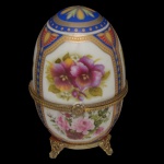Limoges. França - Caixa decorativa em porcelana francesa da manufatura Limoges. Apresenta formato de um ovo com dobradiça central para abertura sustentado por pequenos pés dourados. Pintura a mão de flores (violetas e rosas_ com bordas azuis e detalhes dourado. Marca da manufatura no verso.  9 x 6 cm.