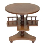 Mesa em madeira nobre, estilo inglês, com tampo circular liso. Coluna central reta e anelada, sustentando à meia altura 4 pequenas prateleiras para livros, bordadas por delicados balaústres. Plateau inferior de formato triangular apoiado em pés baixos. 62 x 60 cm. Sinais de uso e do tempo, com pequenos bicados nas quinas.