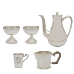 Lote constando de 5 peças em metal prateado sendo: 2 taças baixas de formato circular (8 x 9 cm), B) Bule em metal prateado da manufatura nacional Wolf (18 x 16 cm). C) Caneca em metal prateado e liso. Reserva central (7 x 8 cm) e D) Cremeira em metal prateado. Cabo em madeira.  (8 x 14 x 8 cm)