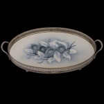 Bandeja de formato ovalado em cerâmica. Borda vazado e alças em metal prateado. Centro com decoração floral na cor azul. Marca da manufatura no verso. 4 x 40 x 27 cm.