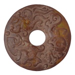 Antigo e raro Disco Bi, artefato ritual chinês, em jade mesclado em tons de marrons e verdes, de formato circular com centro vazado. Apresenta decoração esculpida em alto relevo com dragão e fênix, e verso com pictogramas gravados. Possíveis infiltrações de outros minerais. 12 cm.