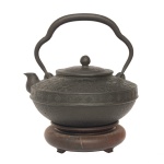Tetsubin", chaleira japonesa em ferro, de formato globular achatado, finamente martelada até meia altura e encimada por friso saliente, faixa em gregas e parte superior retraída, com adornos florais estilizados. Alça superior móvel, recortada em arcos. Bico com extremidade recurva. Tampa levemente abobadada, seguindo a decoração, com pega em pinha. Base baixa e arredondada. Assinada pelo mestre Hojudo ou Hojyu-Do (1755- 1831), com 3 caracteres em relevo na parte superior. Final do séc. XVIII/Início do séc. XIX. Acompanha base em madeira. 18 cm (MT com a alça). 11.5 cm (sem a alça). 17 cm (diam).