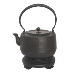 Grande Tetsubin", chaleira japonesa em ferro, de formato cilíndrico reto com parte inferior levemente retraída. Apresenta decoração em leve relevo, com finos filetes salientes e galhos de cerejeiras em flor com parte de cercado. Alça superior circular e bico com extremidade recurva. Tampa plana, levemente rebaixada, em bronze decorado com manchas em laca de cor vinho, característica das tetsubin feitas em Nara. Pega em bolota gravada. Base baixa e arredondada. Início do séc. XIX. Acompanha base em madeira. 22.5 cm (MT com a alça). 14.5 cm (sem a alça). 18.5 cm (diam).