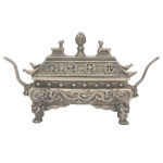 Incensório sino-tibetano para varetas de incenso, de formato retangular em metal prateado usado em altares. Bojo apoiado em patas com garras, sustentando paredes laterais ornadas em relevo com dragões. Pegas laterais no feitio de cabeças de touro com longos chifres. Tampa com lados inclinados, realçados por medalhões e pequenas pegas com pássaros. Parte superior ornada por par de cabras centralizando pinha. Sob a base, símbolo ritual budista gravado. 14 x 12 x 6 cm.