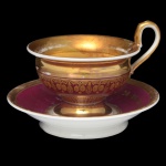 Bela xícara para café com seu pires, em porcelana europeia. Bojo cônico, decorado por faixas com estilizações a ouro sobre fundo vinho, encimado por larga faixa retraída e dourada. Base esparramada seguindo o estilo. Alça lateral em asa. Pires com fundo de cor vinho e borda a ouro liso.8 x 12 cm / 13 cm (pires).