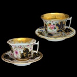 Par de xícaras para café com seus pires, em porcelana europeia policromada e dourada. Bojos abaulados com bordas reviradas, decorados por adornos florais e arranjos com volutas. Interiores dourados. Alças laterais retas. Pratos com bordas lobuladas acompanhando a decoração. 7 x 10 cm / 14 cm (pires).