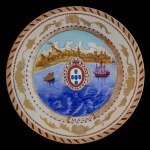 Prato em porcelana Companhia das Índias com temática histórica. Apresenta uma Vista da cidade de Macau, com paisagem costeira e embarcações. No centro o Brasão de Armas de Portugal com a inscrição Macao na parte inferior.