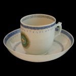 Xícara com seu pires em porcelana, apresenta um emblema central circular com tons de verde, vermelho e dourado, rodeado por um padrão geométrico no topo, Borda com frisos na cor azul.  7 x 7 cm (xícara). 14 cm (pires).