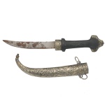 Faca oriental arqueada, com argolas laterais. Bainha e arremates em metal prateado, repuxado e cinzelado, ornados com estilizações e volutas. Cabo em madeira lisa de cor preta, rematada por lâmina em ferro e adornos nas extremidades. (Oxidações na lâmina30 cm.