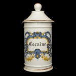 Pote de farmácia em porcelana francesa com filetes dourados, de formato cilíndrico, realçada por anelados em relevo. Apresenta rótulo policromado centralizando o nome do produto COCAÏNE. Tampa cônica com borda saliente, encimada por pega em bolota. Sob a base, marca da manufatura D T Paris. 27 x 12 cm.