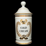 LIMOGES - Pote de farmácia em porcelana francesa com filetes dourados, de formato cilíndrico, realçada por anelados em relevo. Apresenta rótulo em tonalidades douradas centralizando o nome do produto COLD CREAM. Tampa cônica com borda saliente, encimada por pega em bolota. Sob a base, marca da manufatura Au Pot de Galien em Limoges. 28 x 11 cm.