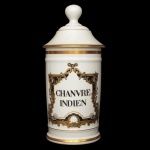 LIMOGES - Pote de farmácia em porcelana francesa com filetes dourados, de formato cilíndrico, realçada por anelados em relevo. Apresenta rótulo em tonalidades douradas centralizando o nome do produto CHANVRE INDIEN. Tampa cônica com borda saliente, encimada por pega em bolota. Sob a base, marca da manufatura Au Pot de Galien em Limoges. 26 x 11 cm.