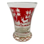 Copo em grosso cristal da Boêmia, com bojo cônico e laterais levemente reviradas. Apresenta frente com reserva em overlay vermelho, ornamentada por paisagem florestal com cervo e árvores. Base saliente, circular e anelada, decorada por treliça de mesma cor. 12 cm