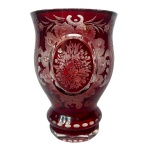 Copo em grosso cristal da Boêmia, de formato abaulado, revestido por overlay de cor bordô. Bojo com laterais recurvas e borda revirada. Apresenta frente com exuberante decoração. Exibe arranjo floral em medalhão oval, emoldurado por composição vegetal realçada por volutas e lavrada em suaves degradês. Base saliente com anelado duplo e facetado, ornado por pequenas janelas lapidadas. 13.7 cm.